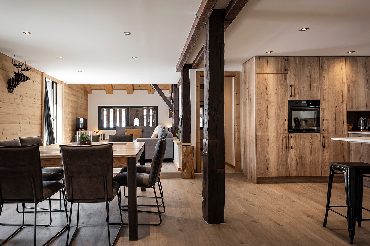 An open living space features a dining area with a wooden table surrounded by six chairs, and a modern kitchen with wood cabinetry and built-in appliances. Light floods in through large windows and a cozy seating area is visible in the background, inviting relaxation.
