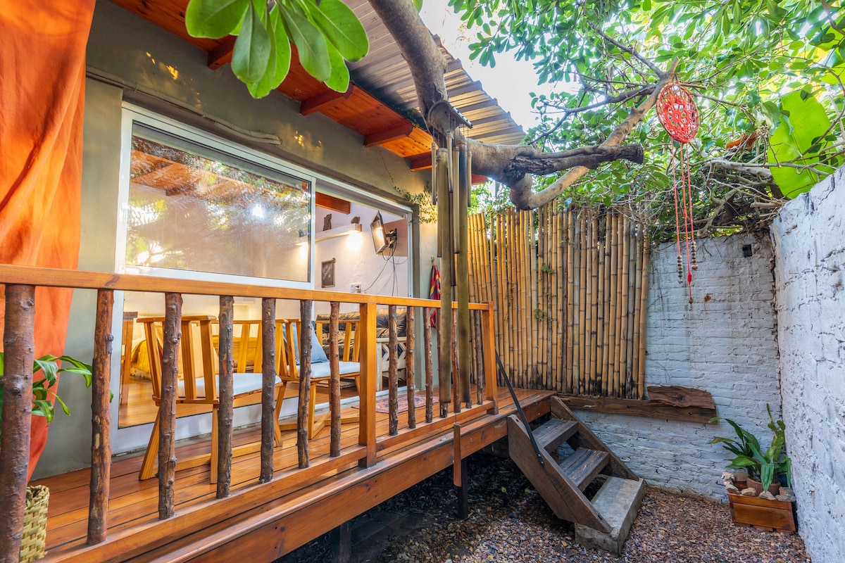 An outdoor deck made of natural wood features rustic railings and steps leading to a small garden area. Lush greenery surrounds the space, enhancing the tranquil environment. The entrance is visible through large glass doors, allowing for ample natural light and views of the serene setting.