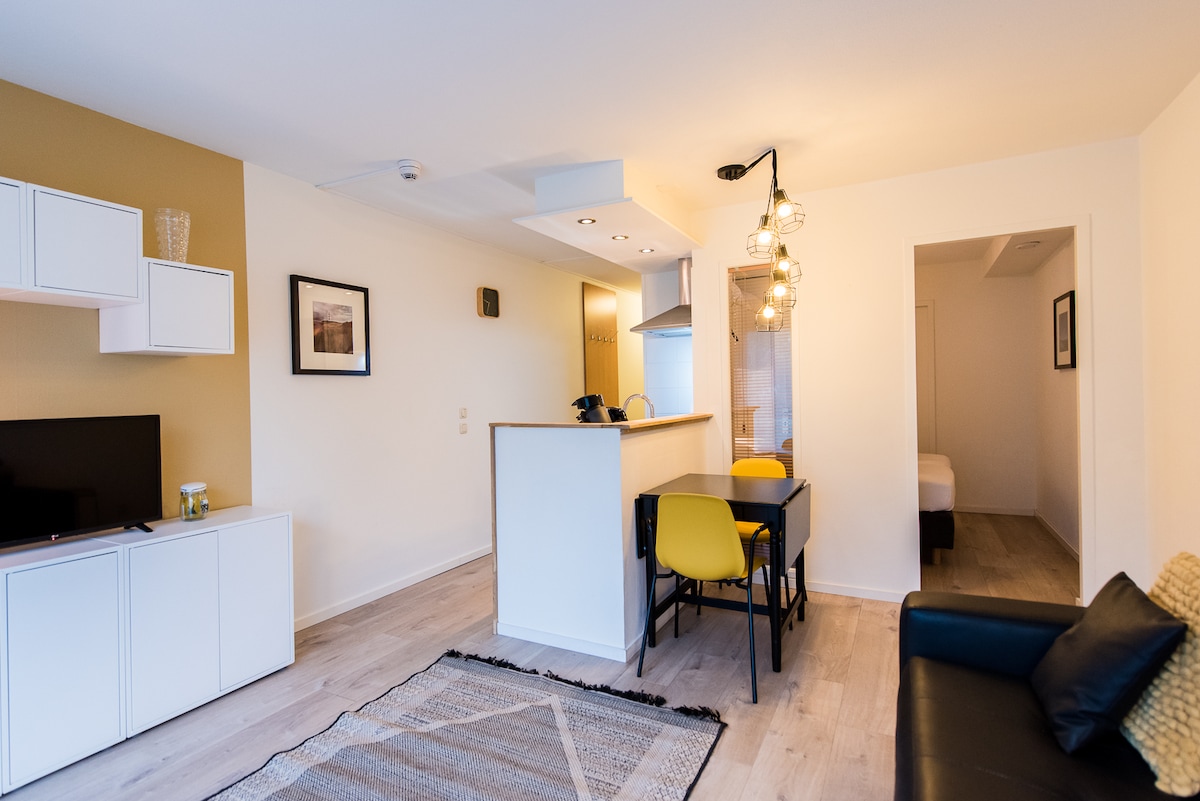 The living space features a combination of a sleek television unit and a minimalist dining area with yellow chairs. An open kitchen is visible in the background, complemented by modern lighting. A subtle area rug lies beneath a cozy couch, enhancing the inviting ambiance.