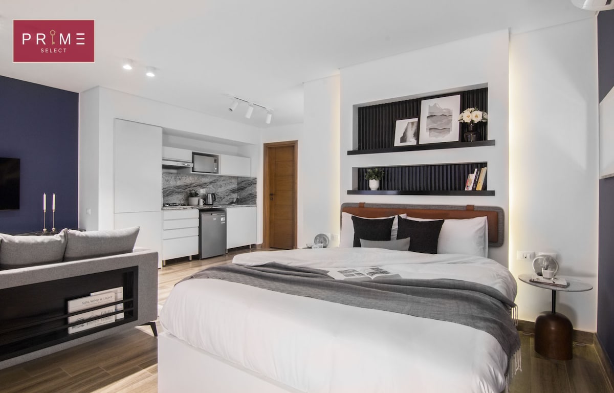 A modern sleeping area features a spacious bed with a neatly arranged gray and white bedding. The room is complemented by a sleek kitchenette and a comfortable seating area, with warm lighting enhancing the contemporary decor. Tasteful artwork is displayed on the walls.