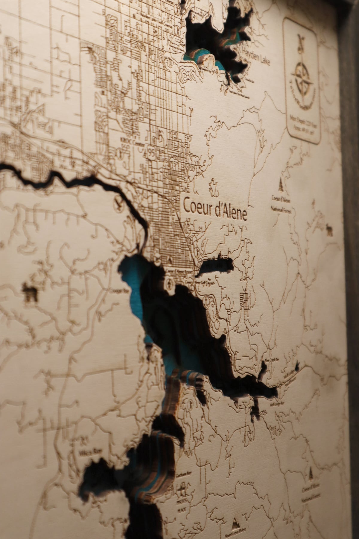 A detailed wooden map of Coeur d'Alene is showcased on the wall. The layers create a three-dimensional effect, highlighting the contours of the lakes and surrounding areas. The design features engraved text labeling specific locations, enhancing the geographical appreciation of the area.