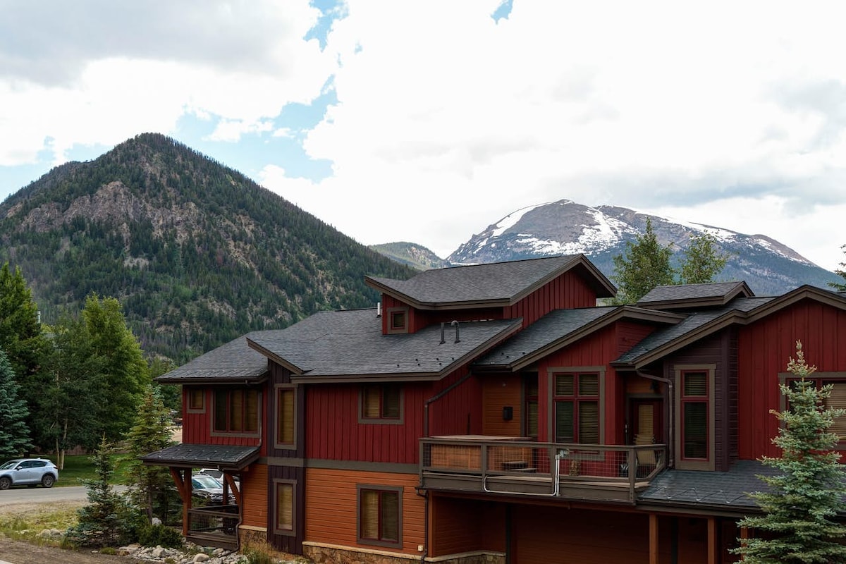 A two-story private home is situated against a backdrop of majestic mountains. The exterior features a combination of deep red and natural wood finishes. Surrounding trees and landscaped details enhance the home's natural appeal, while a balcony offers a vantage point for scenic views.