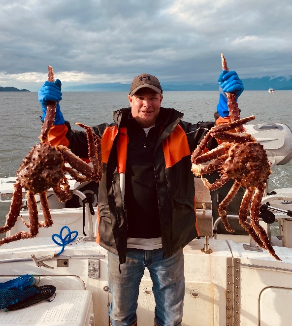 A person holds two large crabs above their head, wearing blue gloves and a jacket. The backdrop features an overcast sky and water, suggesting a boating environment. The scene highlights a maritime adventure, with variations in the water's color reflecting the cloudy atmosphere.