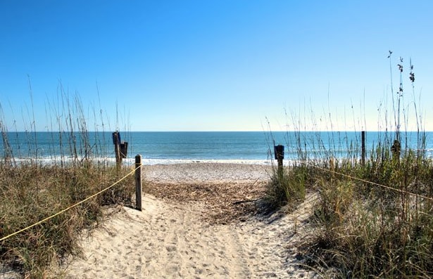 A sandy pathway leads through tall grasses, opening to a serene beach. The calm blue ocean stretches under a clear sky, inviting a sense of peace. Wooden posts and a guiding rope frame the entrance, emphasizing the natural surroundings.
