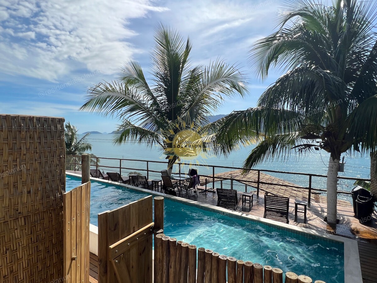 A tranquil outdoor area features a sparkling blue pool surrounded by lounge chairs. Lush palm trees gently sway in the breeze, adding to the serene environment. A clear view of the ocean stretches beyond, with the shoreline visible in the distance.