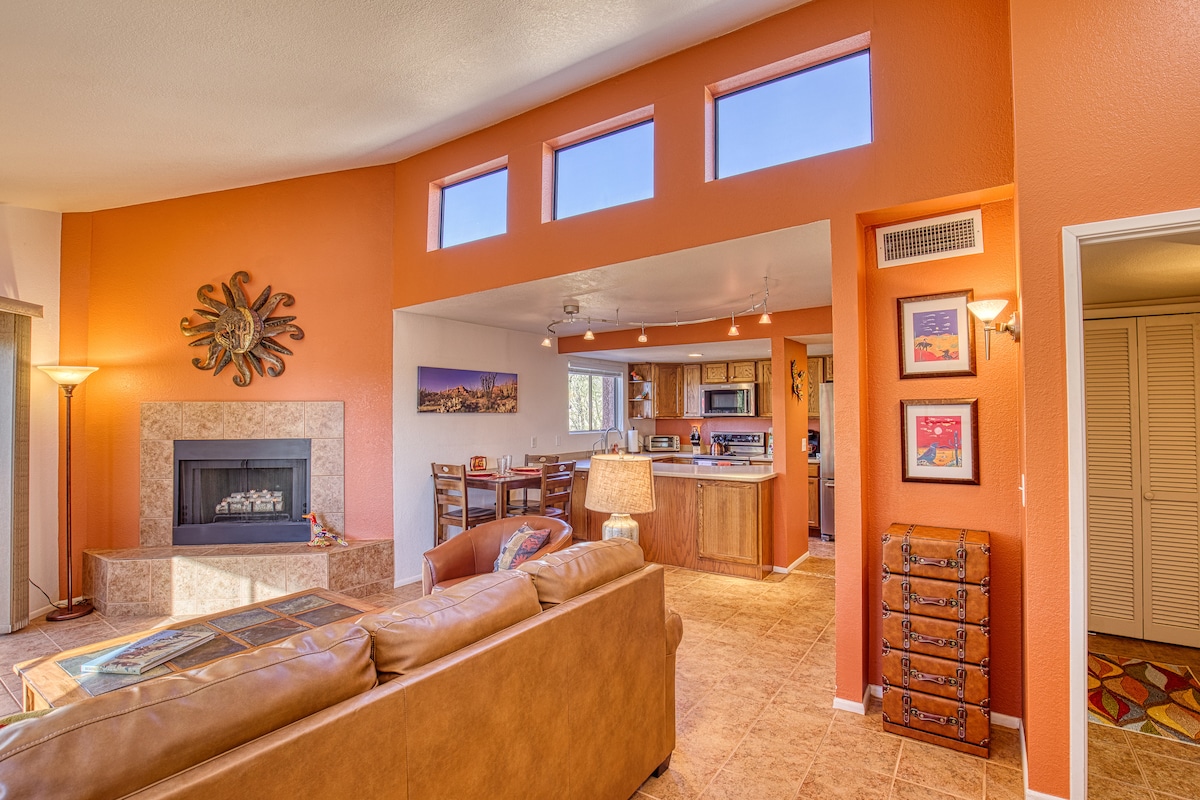 The open-concept living area is characterized by warm orange walls and a cozy fireplace. Natural light floods in through several small windows, enhancing the inviting space. A comfortable couch sits opposite the fireplace, and the kitchen area is visible in the background.