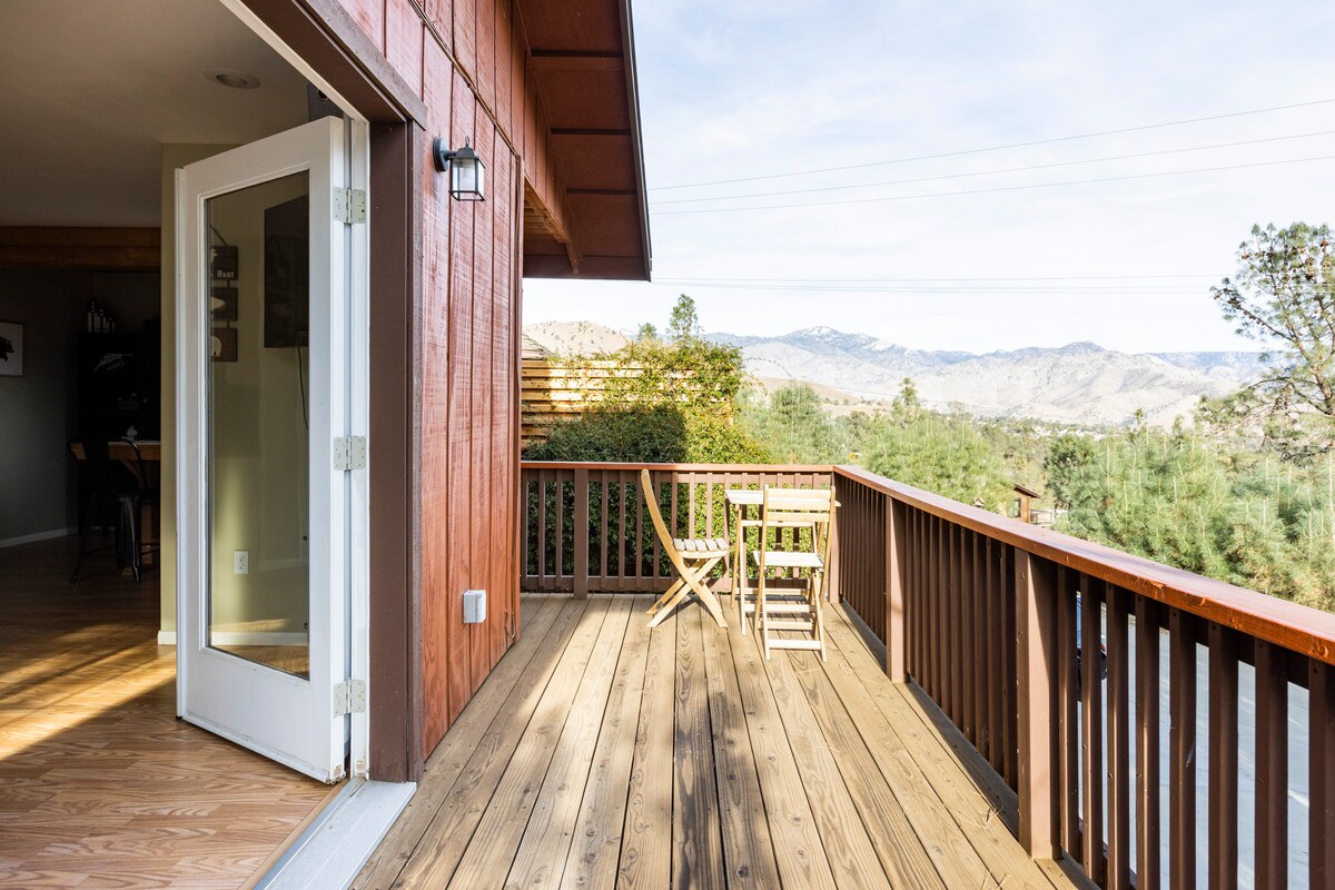 An outdoor balcony features wooden flooring and offers a view of the surrounding mountains. Two folding chairs are positioned invitingly on the deck, with natural light illuminating the space. Large glass doors provide access to the interior of the cabin.