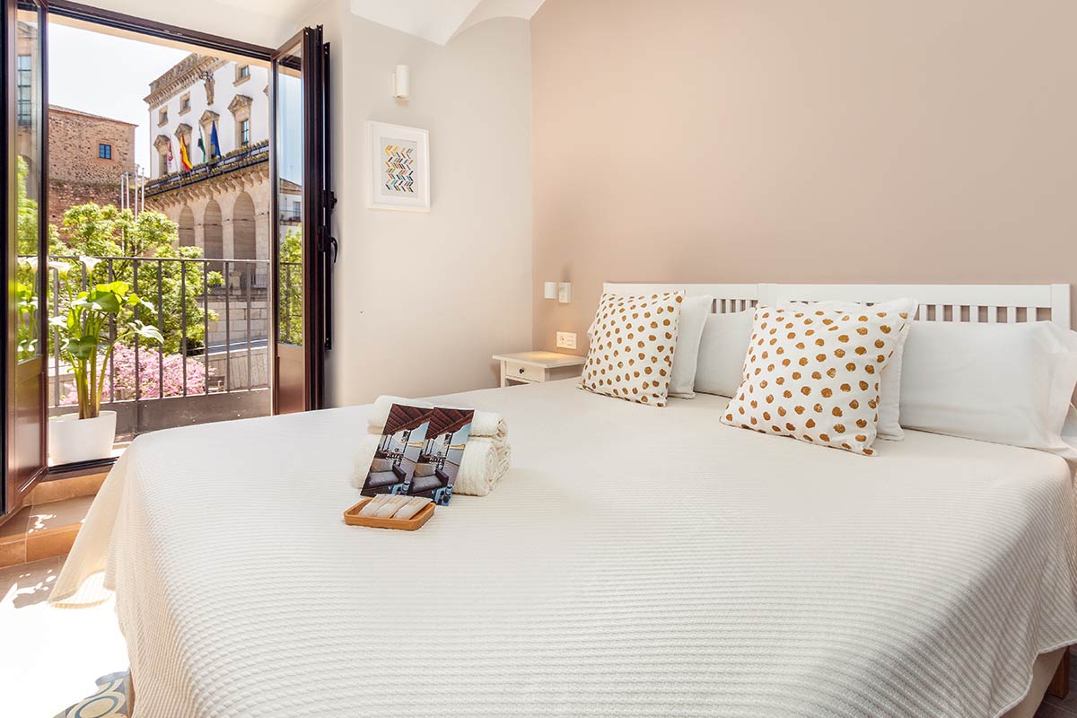 A bright sleeping area features a spacious bed dressed in a light coverlet, flanked by pillows with a subtle pattern. Natural light fills the room through a set of open doors, revealing a view of a vibrant outdoor space with potted plants.