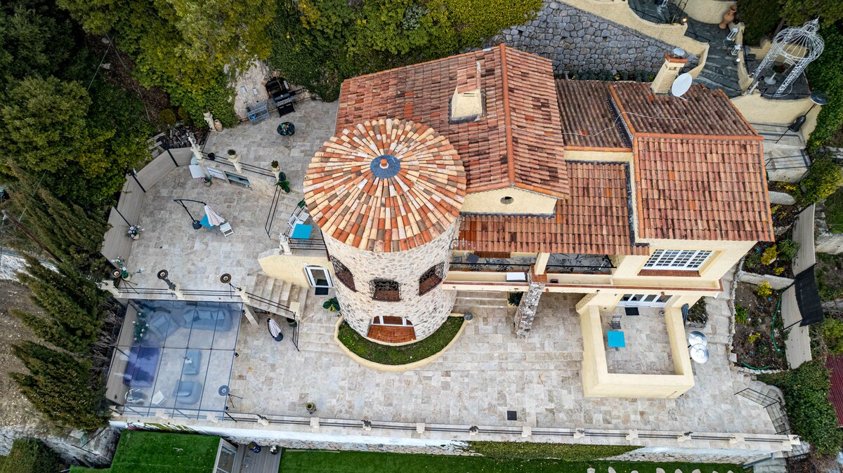 An aerial view of the villa reveals a circular stone tower with a terracotta tile roof. The expansive terrace is lined with lounge chairs and a blue swimming pool. Lush greenery surrounds the area, enhancing the outdoor space's appeal.