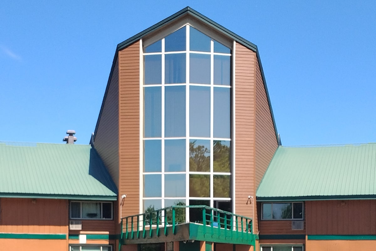 A modern loft condo features a distinctive A-frame architecture with a large central glass window that allows natural light to fill the interior. The exterior is complemented by a green railing and a sloped metal roof, set against a clear blue sky.