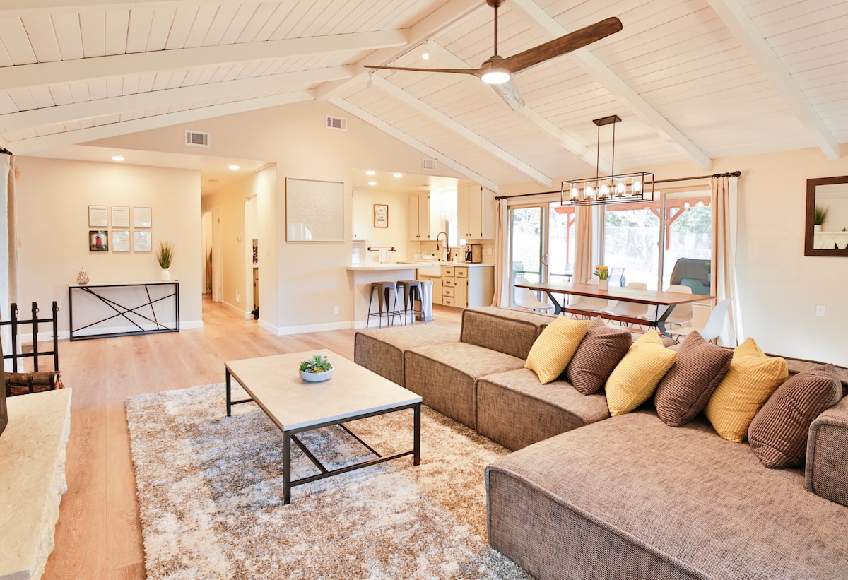 An open-concept living area features a spacious sectional sofa adorned with yellow accent pillows. A sleek coffee table sits on a plush area rug. Vaulted ceilings with exposed beams and modern lighting create an airy feel, while the kitchen and dining area are visible in the background.