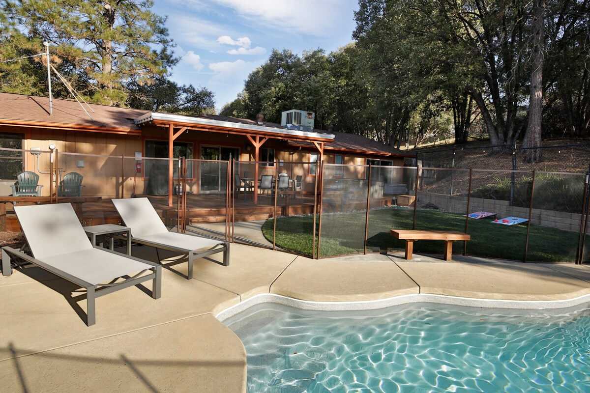 An in-ground swimming pool is surrounded by a deck with two sun loungers. A safety fence encloses the pool area, providing a secure space. The backdrop features a single-story home and lush greenery, while a shaded patio space is visible in the distance.