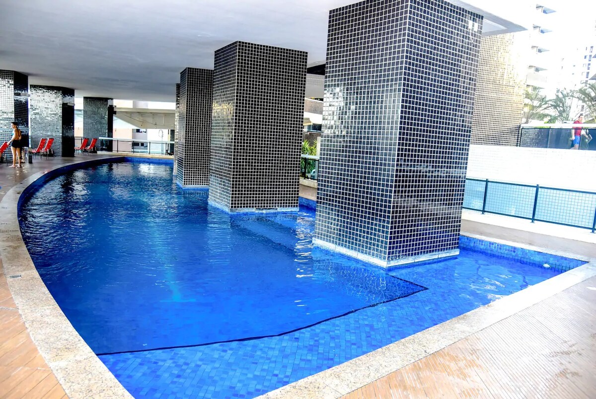 A curved swimming pool is surrounded by tall, tiled columns. The water appears clear, with sunlight reflecting on the surface. Red lounge chairs are positioned nearby, and a portion of the outdoor area is visible in the background, enhancing the overall open space.