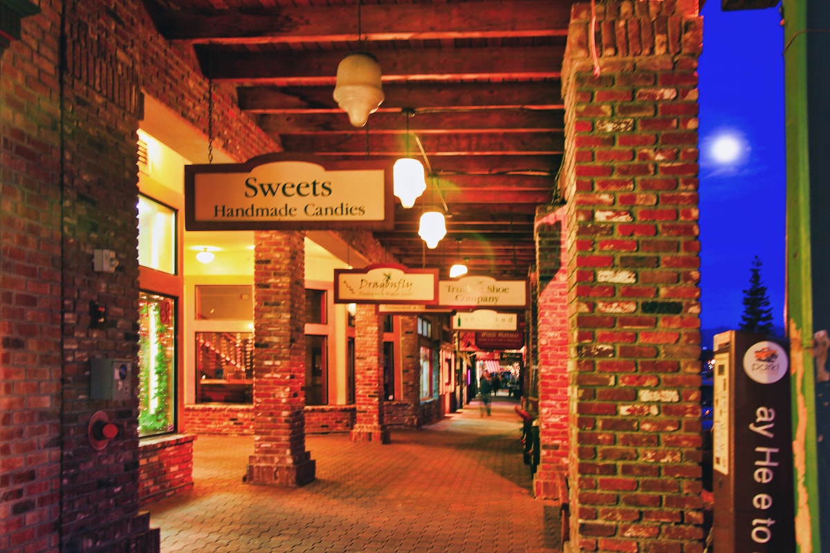 An inviting walkway is lined with brick columns and illuminated by soft lighting. Storefront signs indicate shops including a sweets store offering handmade candies. A partial view of the moon can be seen above, adding to the charm of the evening atmosphere.