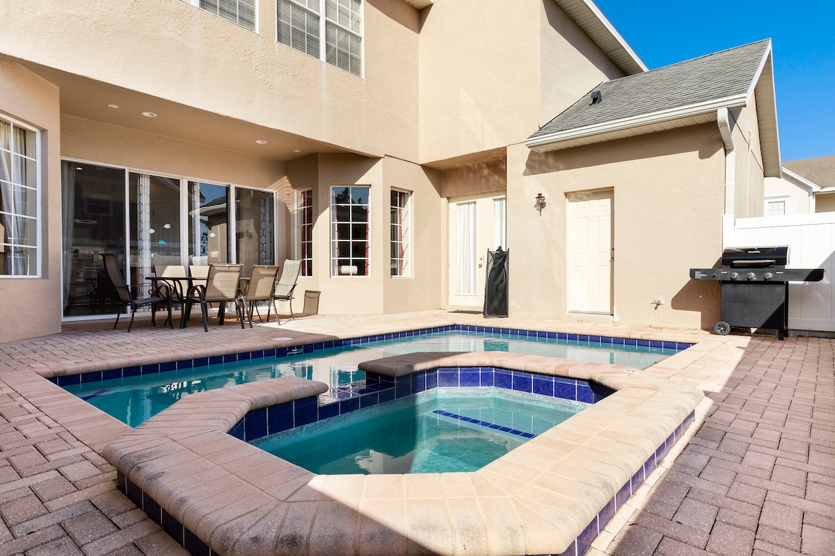 An outdoor area features a private pool and hot tub, surrounded by light-colored pavers. Lounge seating is arranged nearby, with large windows of the house offering views of the space. A barbecue grill is situated at the edge of the deck.