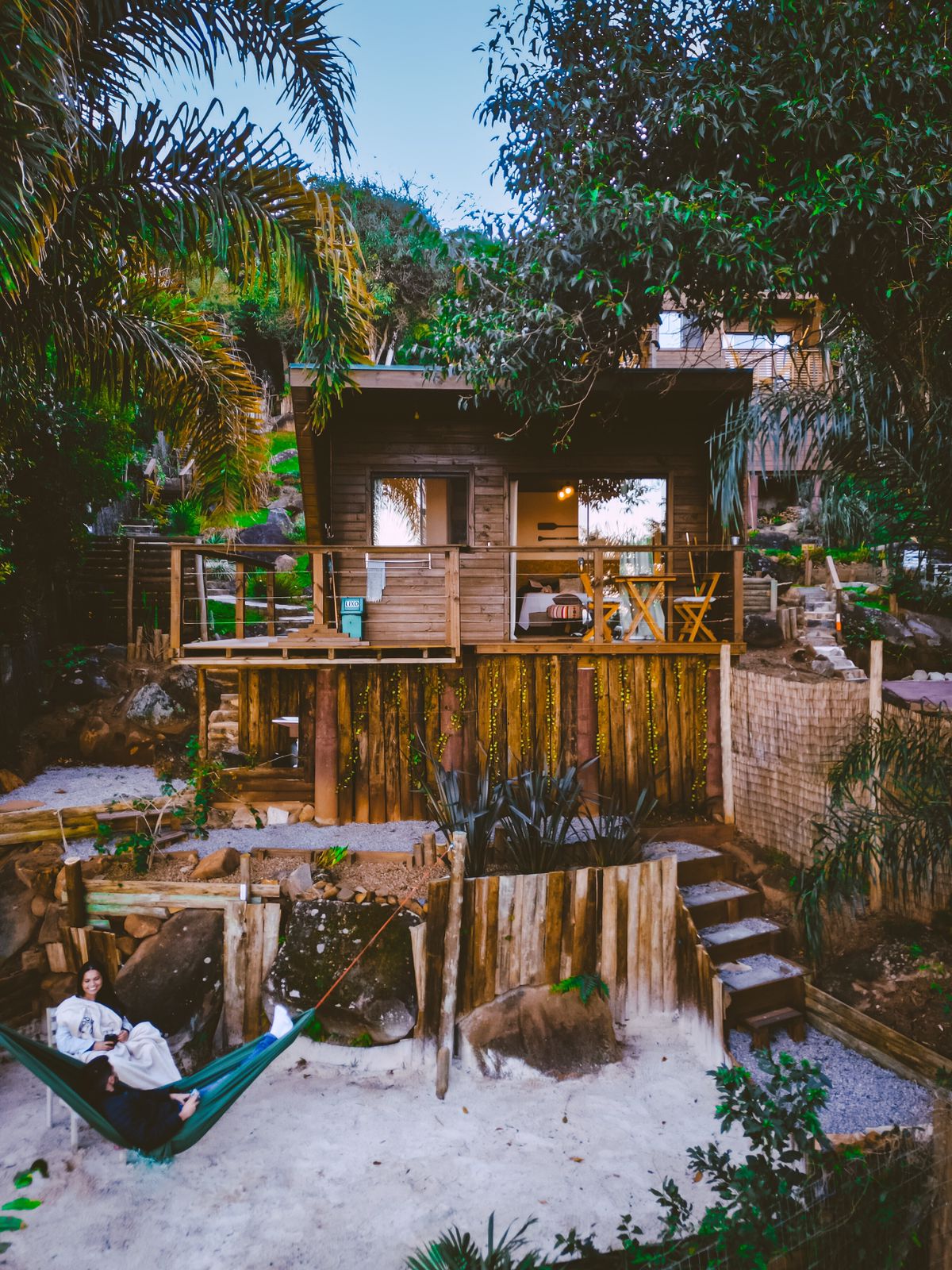 A wooden structure elevated on stilts is surrounded by lush greenery. A small deck with two chairs invites relaxation, while a hammock is nestled below. Steps lined with stones lead to a landscaped area featuring rocks and plants.