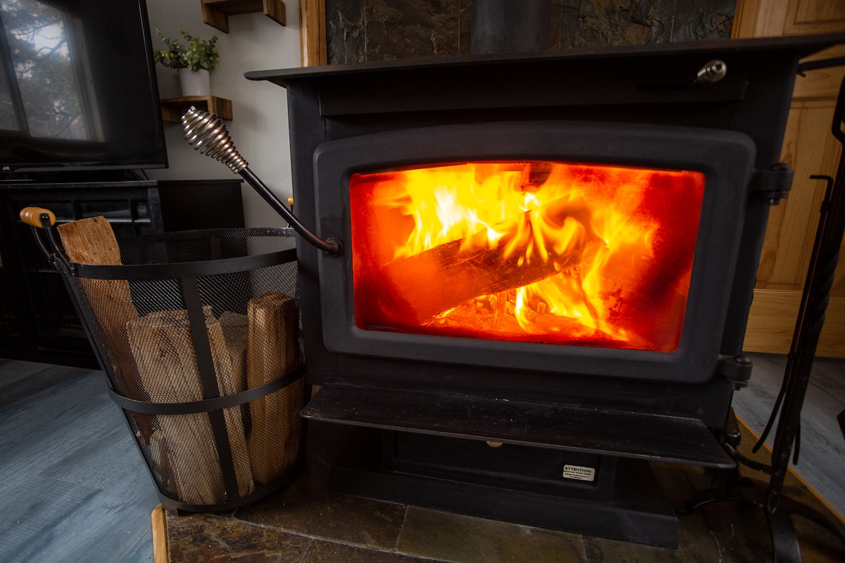 A black wood-burning stove is showcased, featuring a bright orange flame burning inside. Next to the stove, a wire basket containing logs is visible, providing a convenient source of firewood. The surrounding area highlights a cozy and inviting setting.