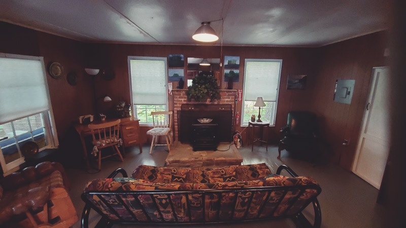 The open-concept living room features a comfortable sofa with a patterned fabric, facing a rustic fireplace adorned with greenery. A wooden dining table is positioned nearby, accompanied by various seating options. Light filters through large windows, enhancing the cozy feel of the space.