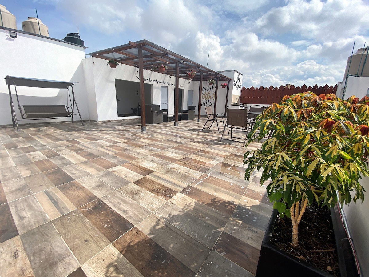 A spacious outdoor terrace is depicted, featuring a tiled floor and a pergola structure. Comfortable seating arrangements are available, along with a swing. Planters with greenery add a touch of nature to the area, which is bathed in natural light.