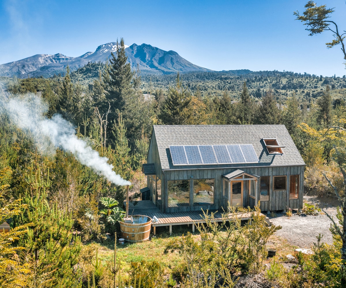 A rustic cabin features large windows and solar panels on the roof, surrounded by native forest. A plume of smoke rises from a nearby outdoor bath, contributing to the tranquil atmosphere. In the background, the volcanic landscape of Calbuco is visible.