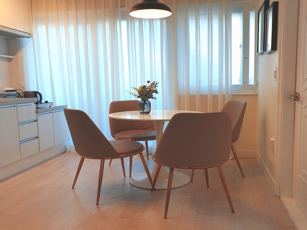 A dining area features a round white table surrounded by four light beige chairs with wooden legs. The space is illuminated by a pendant light, and large windows dressed in sheer white curtains allow natural light to filter through.