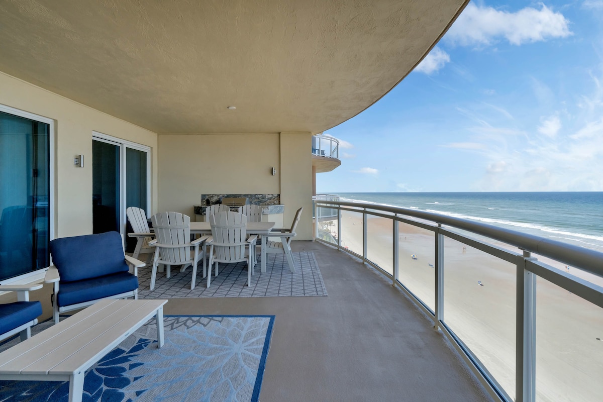 An expansive balcony is furnished with outdoor seating, including a dining area and lounge chairs. The glass-paneled railing provides unobstructed views of the ocean and shoreline, with soft waves visible in the distance. A patterned area rug adds a touch of comfort beneath the seating.