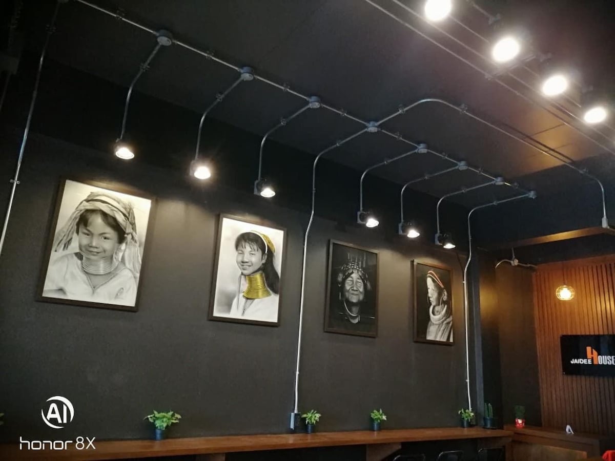 An urban café interior is highlighted by a black wall adorned with framed portraits in monochrome, each missing facial features. Soft industrial lighting hangs from the ceiling above a wooden counter, with small potted plants placed strategically along the surface, contributing to a modern aesthetic.