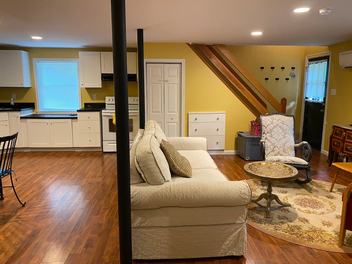 An open living area is showcased with a light-colored sofa positioned beside a coffee table. The kitchen features white cabinets and dark countertops. Natural light enters through a window, illuminating the space which includes wooden flooring and decorative accents.