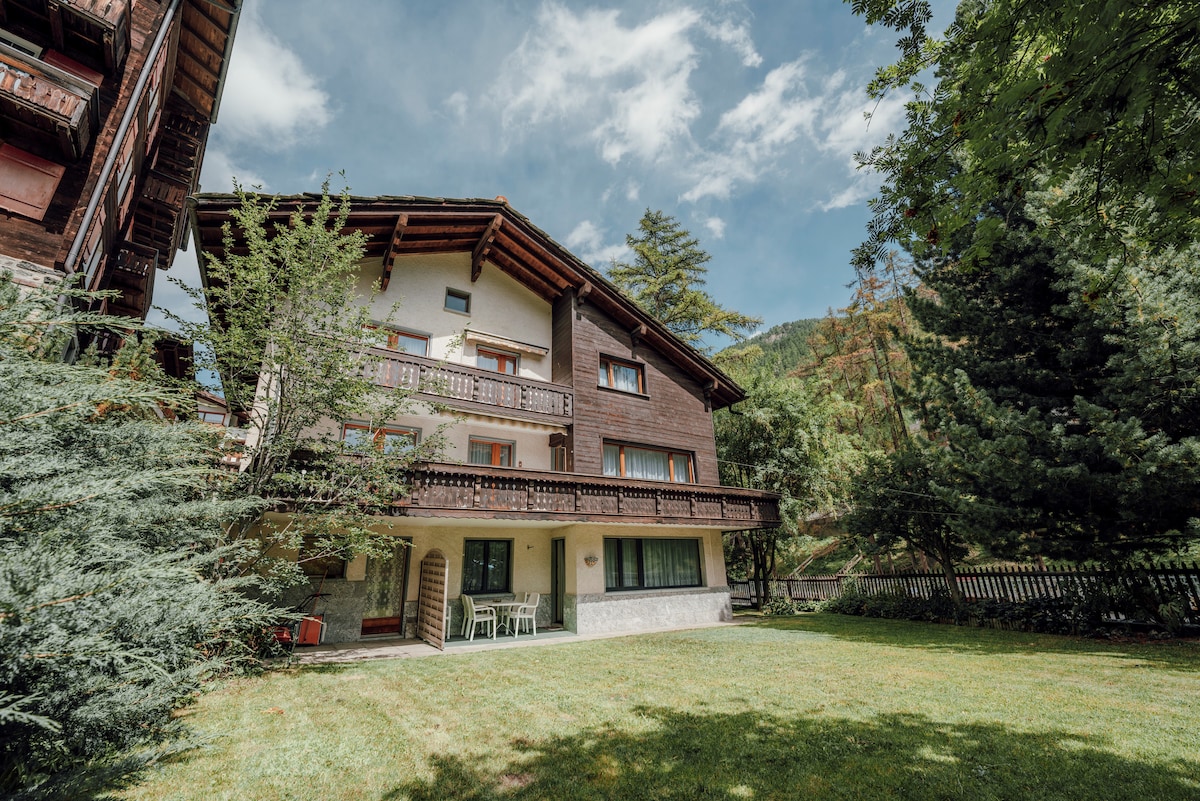 A spacious building with multiple balconies is set amidst lush green grass and trees. The façade showcases wooden elements, and the windows provide a view of the surrounding nature. A small seating arrangement is visible on the patio, contributing to a peaceful outdoor setting.