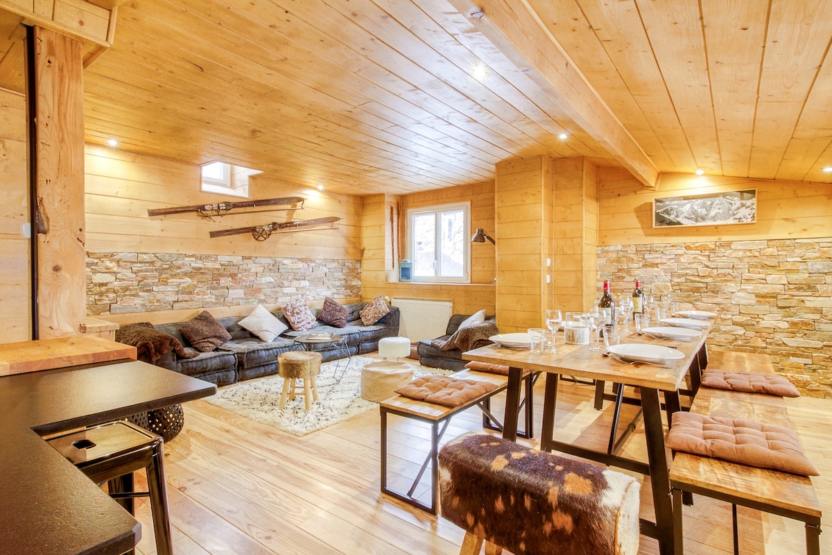 A spacious living area features warm wooden walls and ceiling, complemented by a stone accent wall. A large sectional sofa with assorted pillows invites relaxation. There are multiple dining tables arranged for communal meals, with stylish stools adding to the rustic charm.