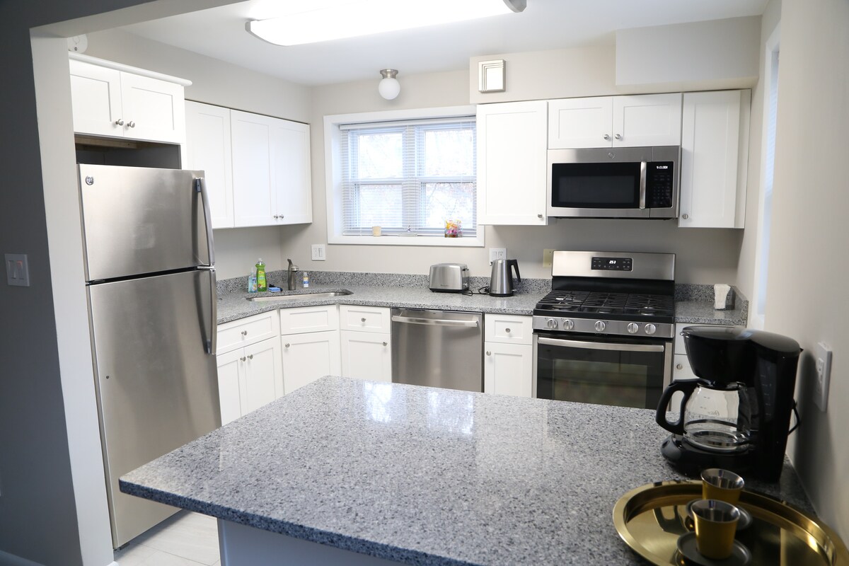 A modern kitchen is showcased, featuring stainless steel appliances including a refrigerator, oven, and microwave. Granite countertops provide ample space. A coffee maker and a tray with cups are neatly arranged on the counter, and natural light enters through the window.