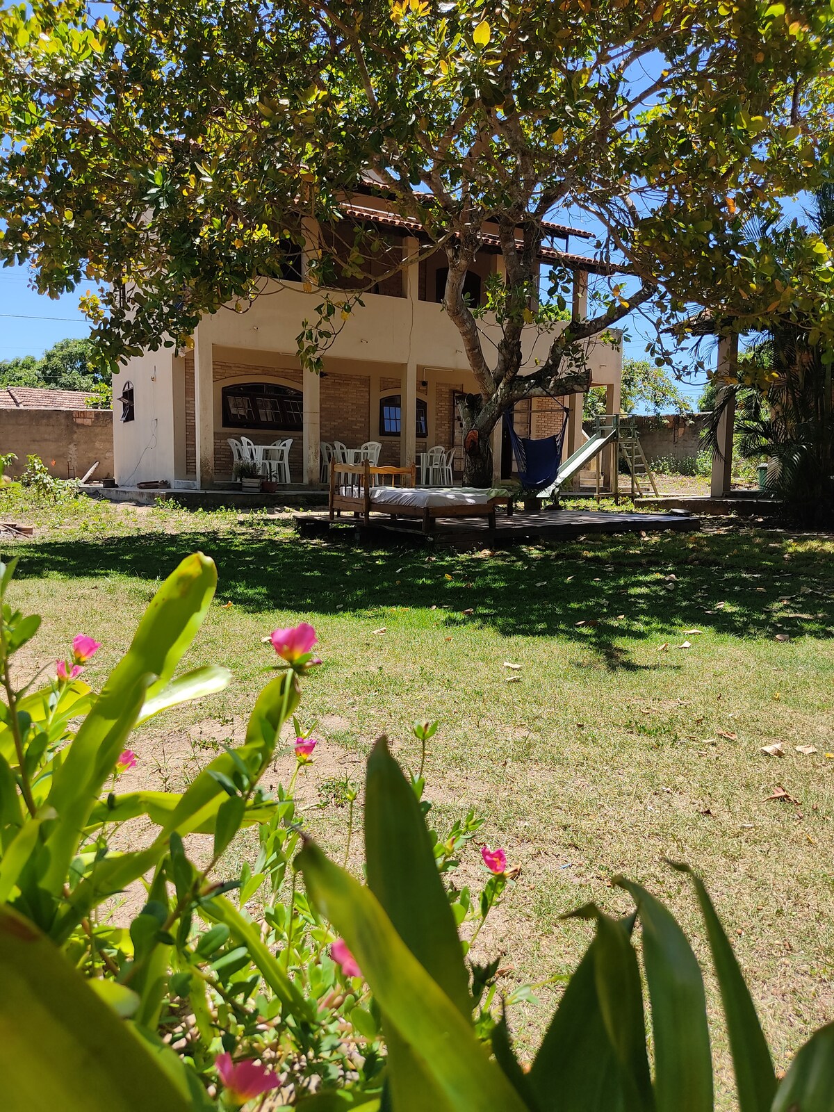 A spacious courtyard is framed by blooming flowers and greenery, with a subtle hint of sunlight casting gentle shadows. The two-story house features a deck with seating areas and a hammock, creating a relaxed outdoor ambiance. The bright sky enhances the inviting atmosphere.