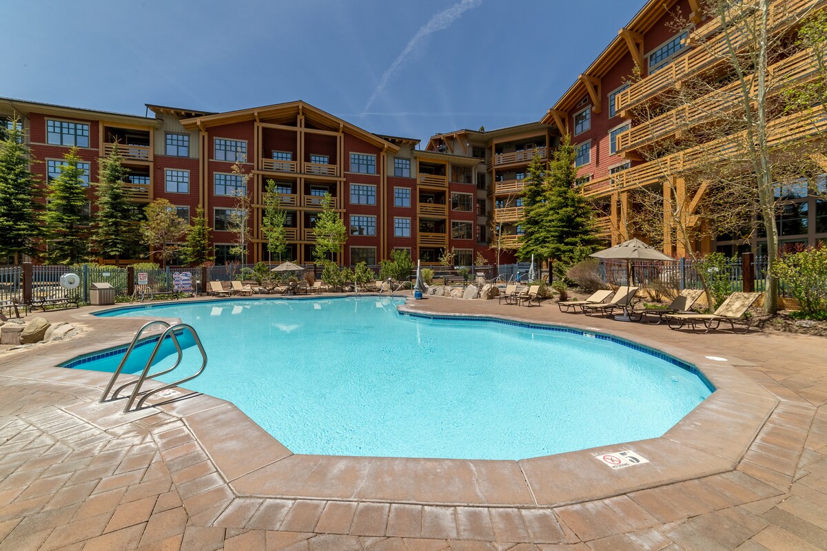 A well-maintained outdoor pool area is featured, surrounded by lounge chairs and umbrellas. Lush landscaping with trees enhances the ambiance. The building's colorful facade and balconies provide a scenic backdrop, inviting relaxation under clear blue skies.