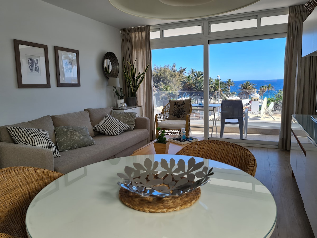 A bright living area features a comfortable sofa adorned with decorative cushions. A round dining table is set with a leaf-shaped centerpiece. Floor-to-ceiling windows provide a view of lush palm trees and the ocean, enhancing the light and airy ambiance.
