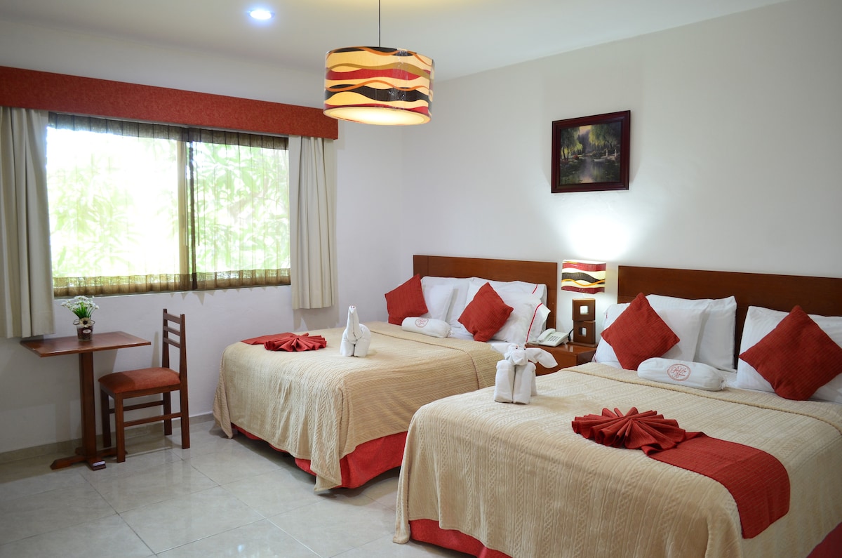 A well-lit guest room features two spacious beds with cream-colored blankets and red accents. A small wooden table and chair are positioned near a large window draped with light-coloured curtains. Decorative towels are arranged on the beds, contributing to the room's welcoming appearance.