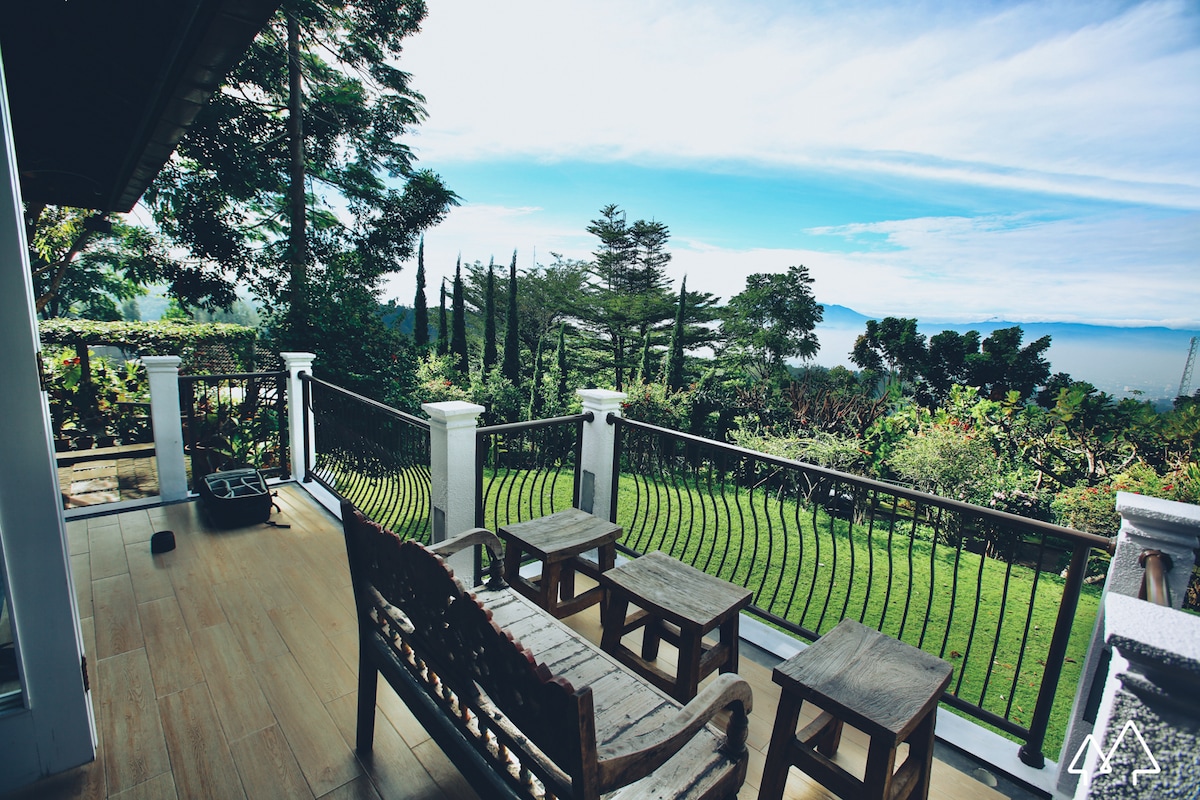A spacious balcony offers a tranquil setting, featuring several wooden chairs arranged around a rustic table. Lush greenery and tall trees can be seen beyond the railing, providing a serene view of the garden and hills in the distance beneath a clear blue sky.