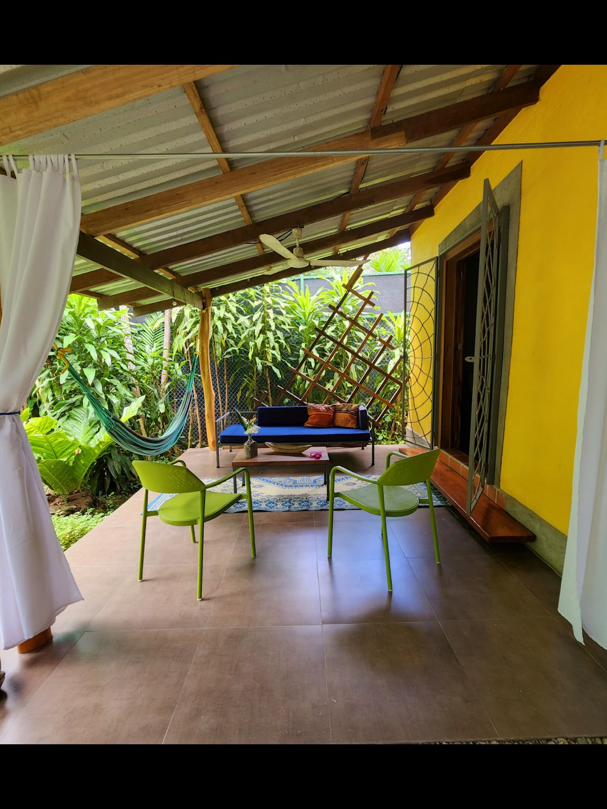 A covered terrace features two green chairs and a coffee table, set against a backdrop of lush greenery. A cozy hammock is visible in the corner, inviting relaxation. The vibrant yellow walls of the studio add warmth to the peaceful outdoor space.