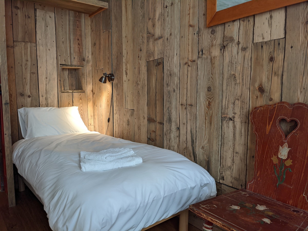 A single bed with crisp white linens is positioned against a backdrop of rustic wooden walls. A small, artistic chair with a heart cutout adds charm to the space, while a reading lamp provides warm illumination, enhancing the cozy ambiance.