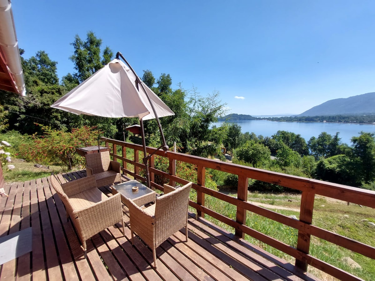 A wooden deck features cozy seating with two woven chairs and a small table, complemented by a large umbrella for shade. Scenic views of the lake and surrounding greenery create a peaceful outdoor space.