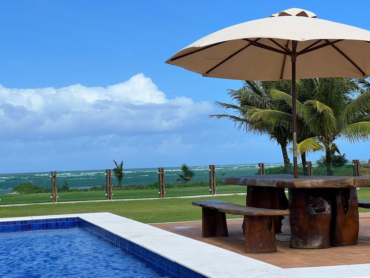 A wooden table with an umbrella is positioned near a swimming pool, offering a view of the ocean in the background. Lush greenery and palm trees surround the area, complementing the expansive blue sky with scattered clouds.