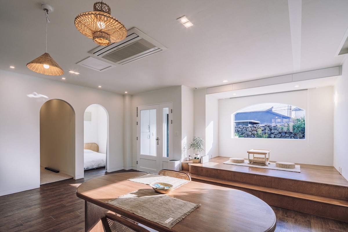 A bright interior space is featured, highlighted by a spacious table and natural wood accents. Arched doorways lead to a serene sleeping area, while large windows allow soft light to fill the room, creating a calm and inviting environment.