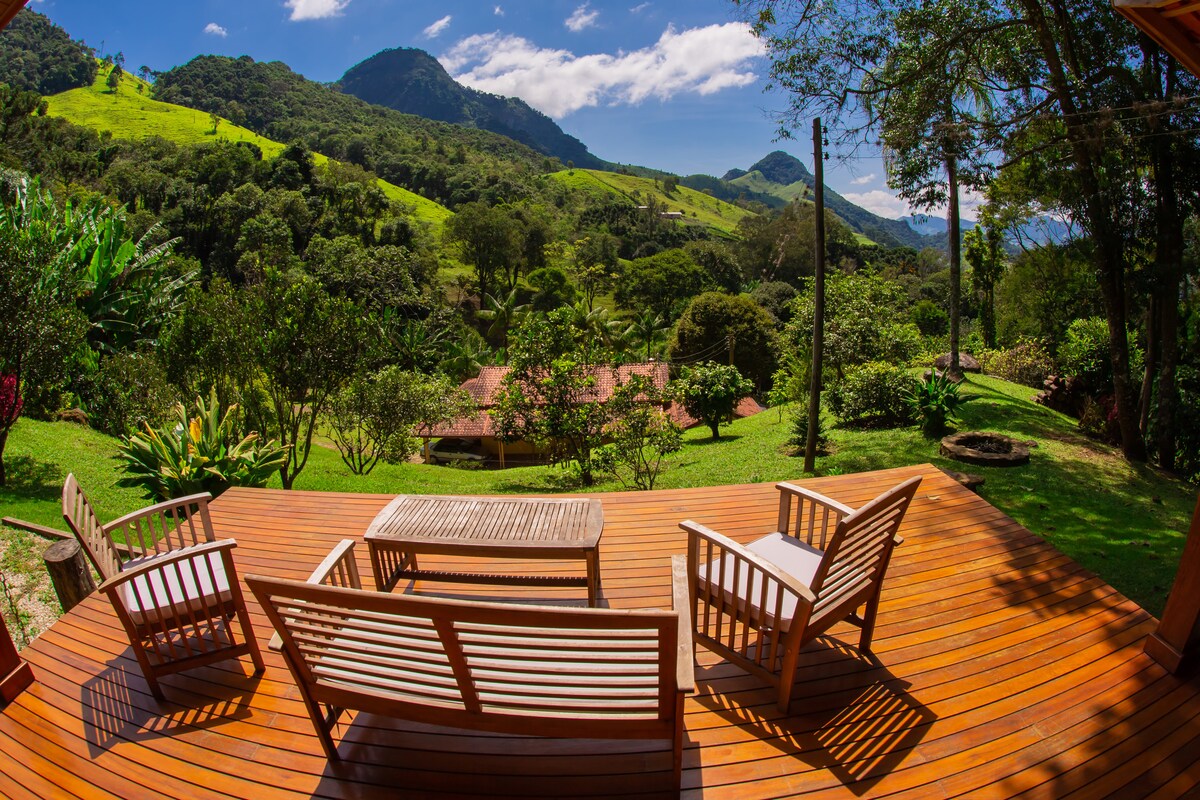 A spacious wooden deck features comfortable seating arrangements, including two armchairs and a table, overlooking lush greenery and rolling hills. The distant mountains are visible under a clear blue sky, offering a serene natural landscape.