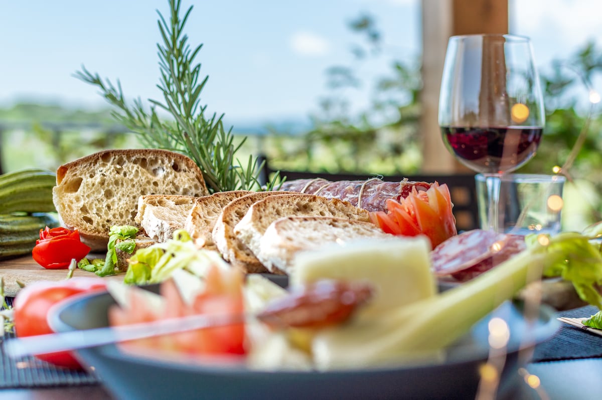 A variety of fresh foods is displayed on a wooden surface, including slices of bread, tomatoes, and cured meats. A glass of red wine sits nearby, and a few sprigs of herbs add a touch of greenery. The background reveals a scenic view of the vineyard.
