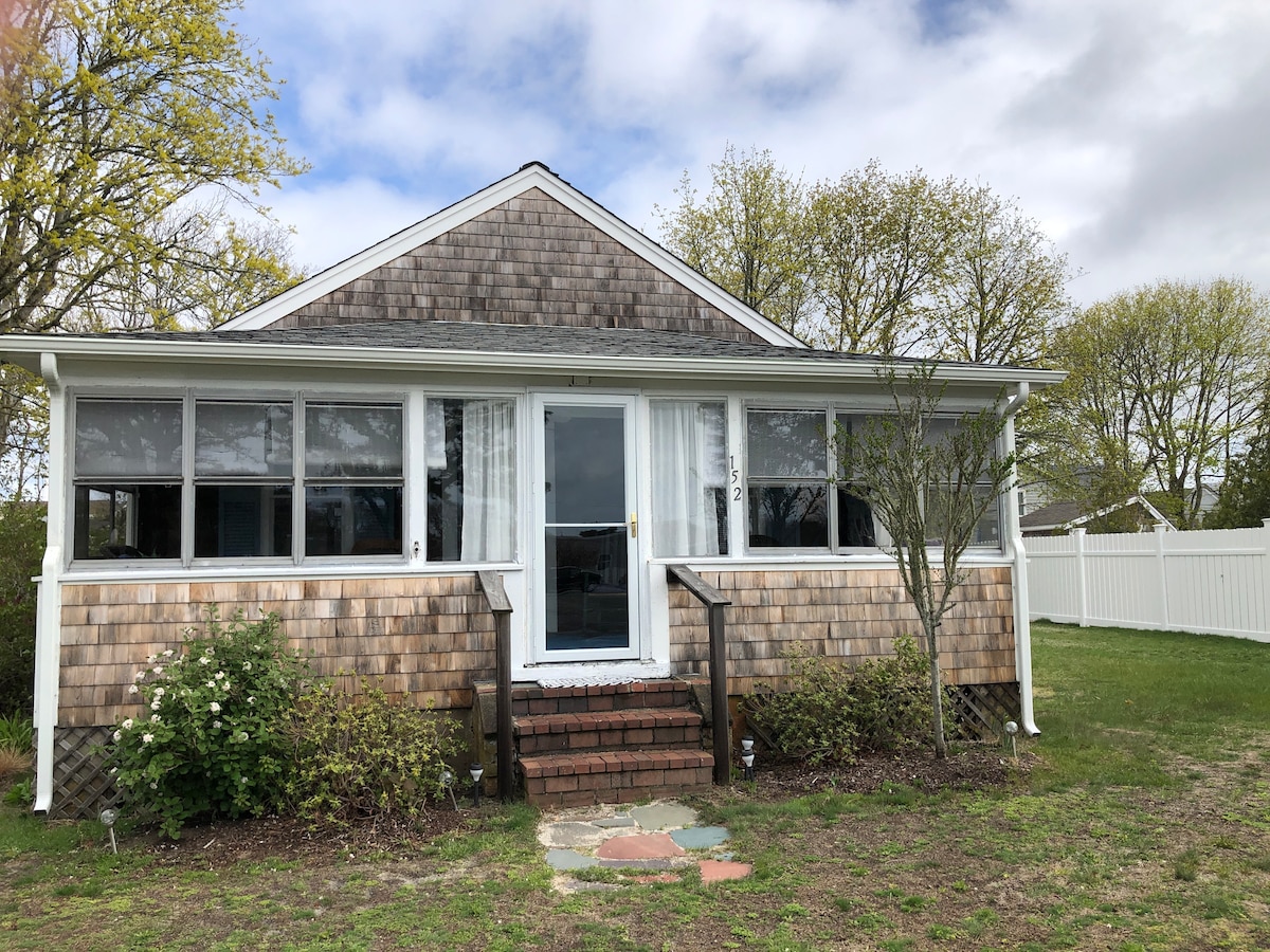 A charming cottage exterior is visible, featuring a shingled facade and large windows that allow for natural light. A set of stone steps leads up to the front door, framed by greenery and small shrubs. The yard is spacious, with trees in the background.