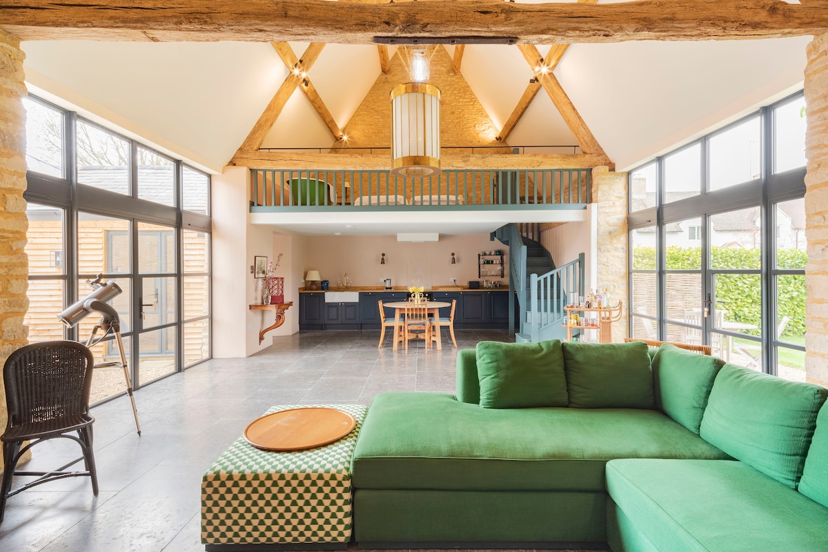 The spacious living area features a large green sectional sofa paired with a patterned ottoman. Sunlight pours in through tall glass doors, illuminating the modern interior. The wooden beam ceiling adds character, and a staircase leads to the mezzanine level, offering access to an open plan kitchen.