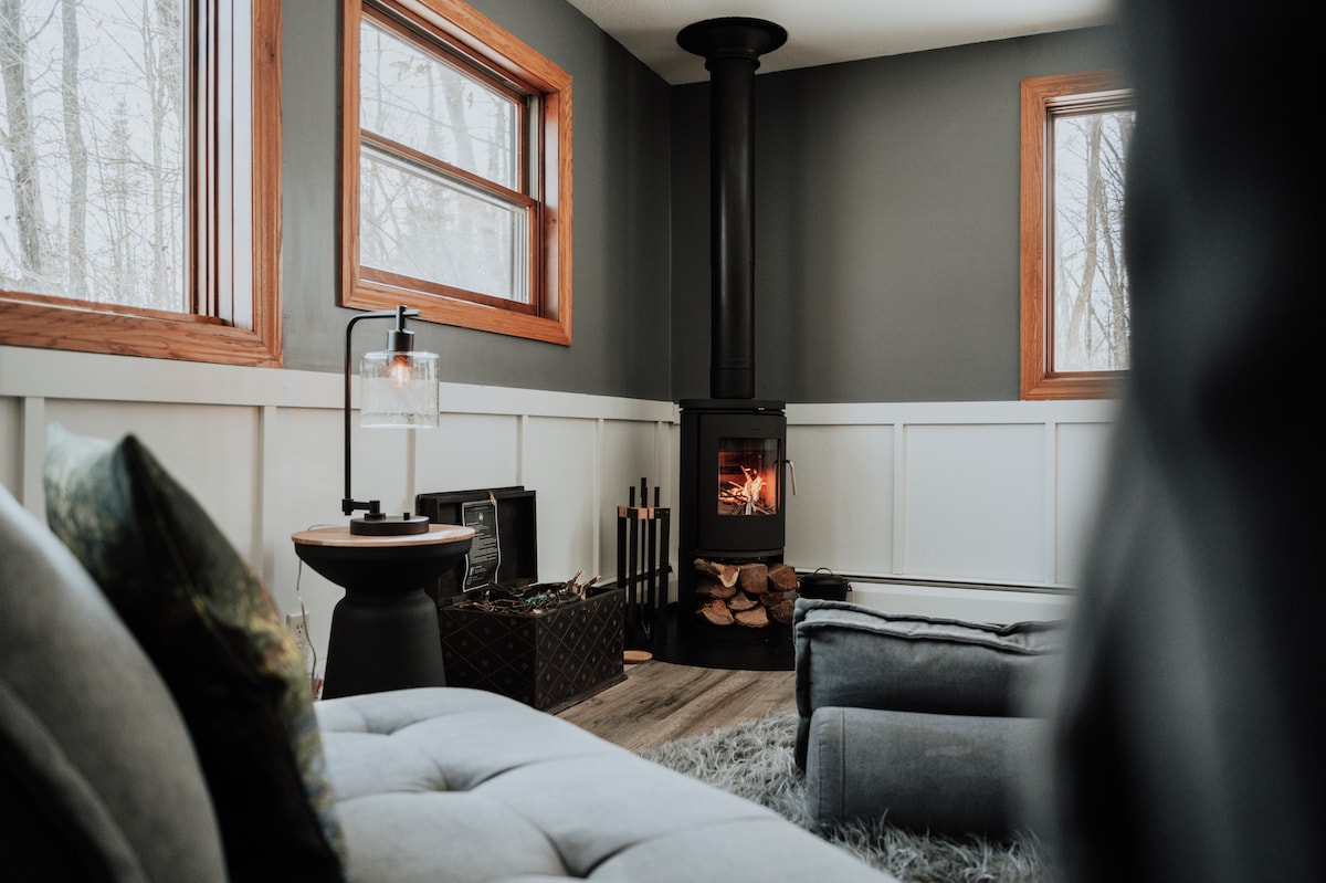 A cozy living room is shown with large windows allowing natural light to fill the space. A wood stove is situated in the corner, contributing to a warm atmosphere. Soft furnishings are arranged for comfort, with a stack of logs nearby for the fire.