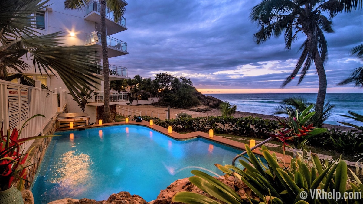 A tranquil beachside pool is framed by tropical plants and palm trees, with the ocean visible in the background. The sky is painted with soft shades of purple and blue as the sun sets, casting gentle reflections on the water's surface.