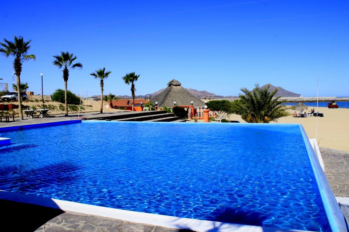 An expansive infinity pool features a vibrant blue surface that reflects the clear sky above. Lush palm trees surround the pool area, leading towards a sandy beach, while a thatched-roof structure and distant hills are visible in the background.