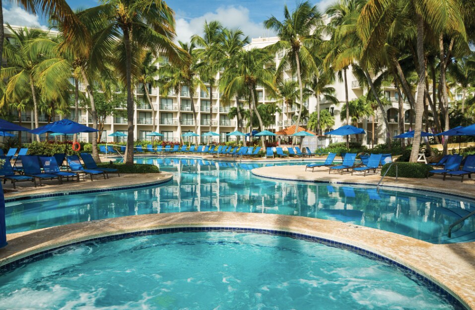 A spacious pool area is bordered by lush palm trees, offering a serene oasis. Lounge chairs in vibrant blue line the water's edge, complemented by shaded umbrellas. The clear water of the pool reflects the sky, creating a tranquil atmosphere for relaxation.