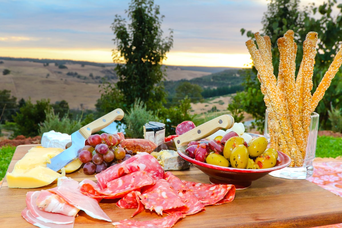 A wooden serving board displays an assortment of charcuterie, including sliced meats, cheeses, and olives, accompanied by fresh grapes. Bright sunlight casts a warm glow over the scene, with distant rolling hills and trees visible in the background.